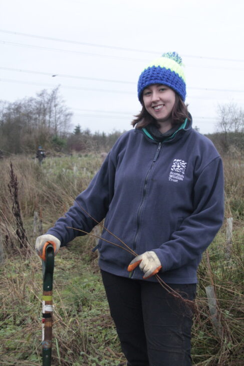 Alex smiling while standing holding a sapling in one hand and a spade in the other